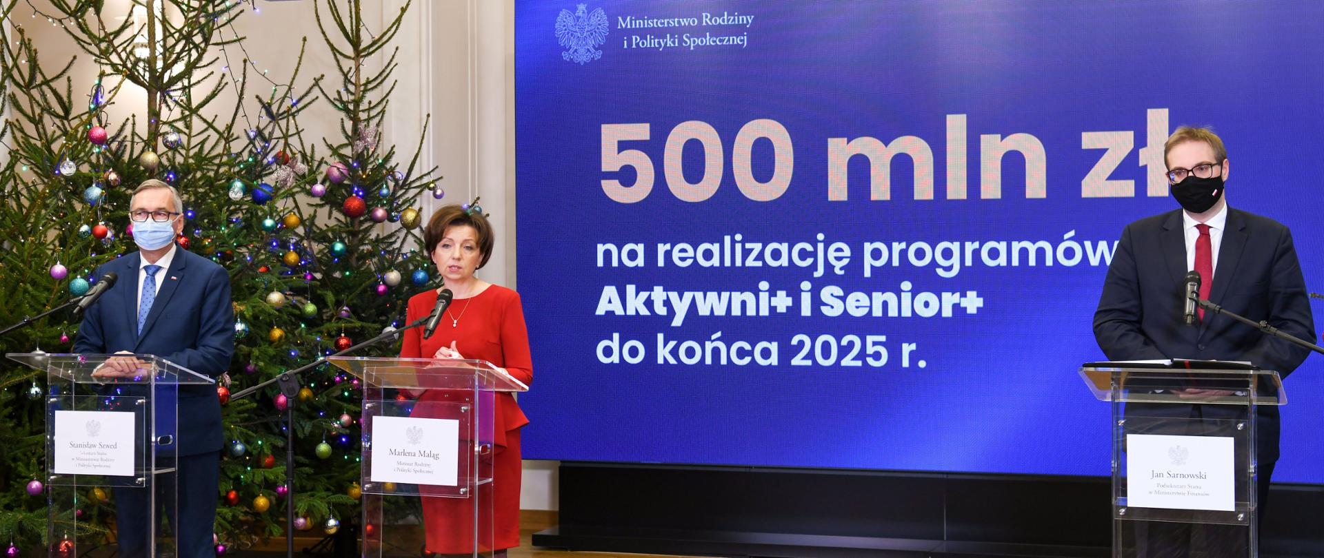 Trzy osoby stoją przy plastikowych, przeźroczystych mównicach. W centralnym punkcie ubrana w czerwony kostium przemawia minister Maląg. W tle baner, na którym na niebieskim tle widoczny jest biały napis 500 mln zł na realizację programów Aktywni+ i Senior+ do końca 2025 r. 