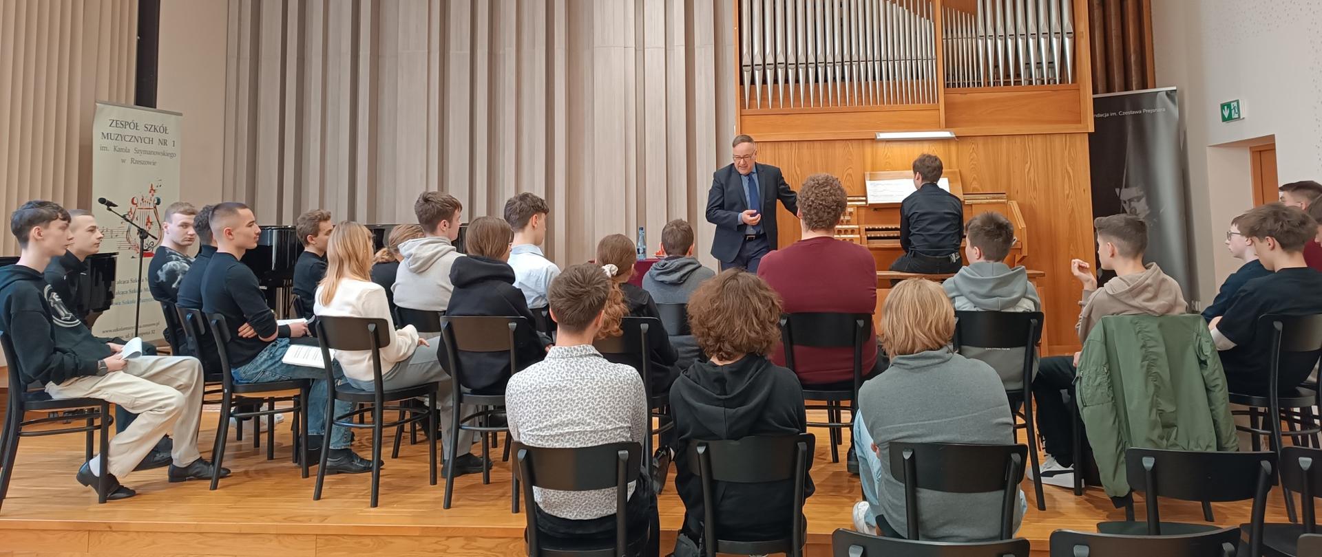 Uczniowie siedzą w sali koncertowej podczas Regionalnych Warsztatów Organowych w Zespole Szkół Muzycznych nr 1 w Rzeszowie, a prowadzący omawia zagadnienia przy organach piszczałkowych.