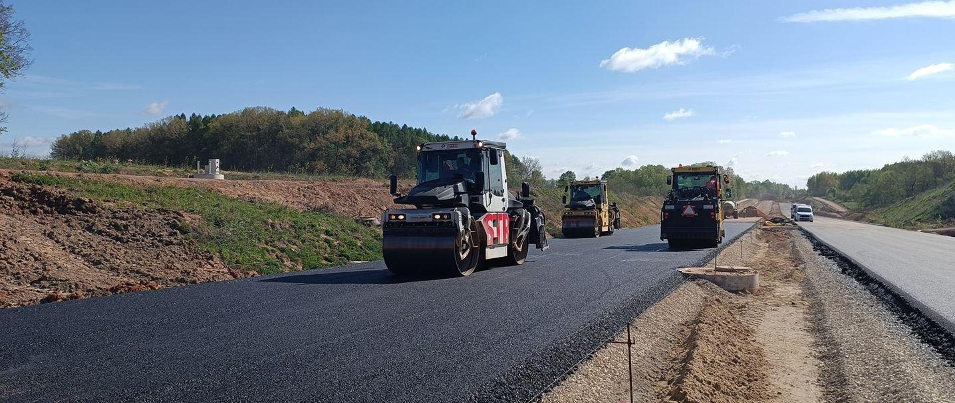 DK42 obwodnica Wąchocka - układanie nawierzchni na budowie dwujezdniowej drogi - trzy walce drogowe jadą po jezdni z nową czarną warstwą podbudowy asfaltowej, po prawej druga jezdnia z szarą warstwą warstwy wiążącej nawierzchni. 