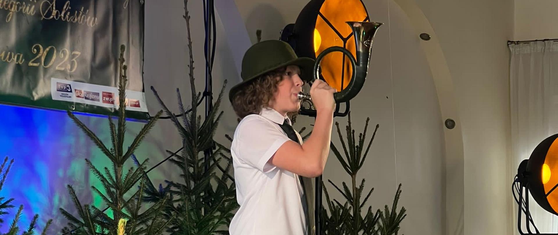 Chłopiec grający na rogu. Scena ozdobiona świerkami. Na ścianie widoczna część ekranu z nazwą imprezy.