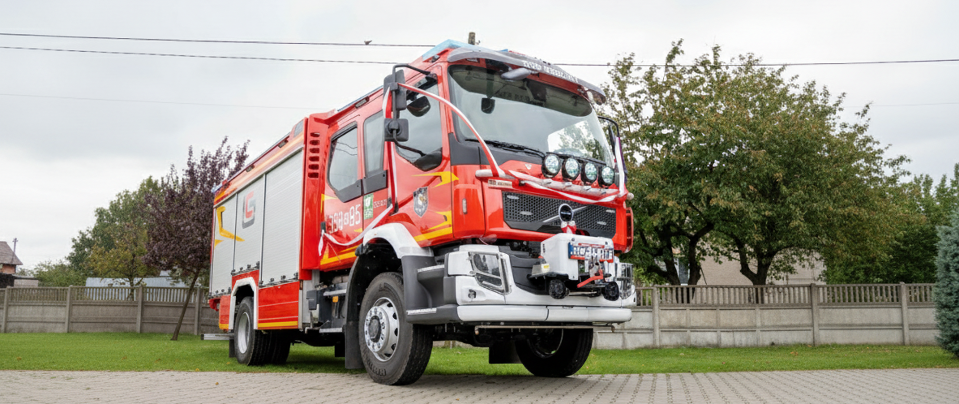 Średnie ujęcie na czerwony wóz strażacki zaparkowany na terenie zielonym, prawdopodobnie przed remizą lub budynkiem użyteczności publicznej. Jest to nowoczesny pojazd pożarniczy. Na szybie przedniej i karoserii widoczne są biało-czerwone szarfy lub wstęgi, co sugeruje, że pojazd został niedawno poświęcony, przekazany lub jest używany podczas uroczystości. Na dachu kabiny kierowcy widnieje napis "OSP REDZINY".
Przód pojazdu to kabina z dużymi, ciemnymi oknami. Na dachu kabiny, nad przednią szybą, znajduje się listwa z dodatkowym oświetleniem. Na atrapie chłodnicy i masce umieszczone są emblematy, w tym herb. Biały zderzak i podwozie kontrastują z intensywną czerwienią reszty nadwozia. Na zderzaku zamontowany jest mały, dodatkowy zraszacz/działko. Na tablicy rejestracyjnej widnieje numer "SLZ 510F".
Boczna część pojazdu to zabudowa z aluminiowymi żaluzjami schowków. Na zabudowie, w kolorze czerwonym z żółtymi detalami, widnieje numer taktyczny "358-85". Koła pojazdu są duże, terenowe.
W tle widać fragment trawnika i ogrodzenia z betonowych płyt, a także drzewa o jesiennych liściach (zielonych i bordowych). Niebo jest jasne, zachmurzone.
