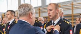 Na zdjęciu znajduje się strażak w mundurze, któremu wręczany jest awans, w tle widać innych strażaków