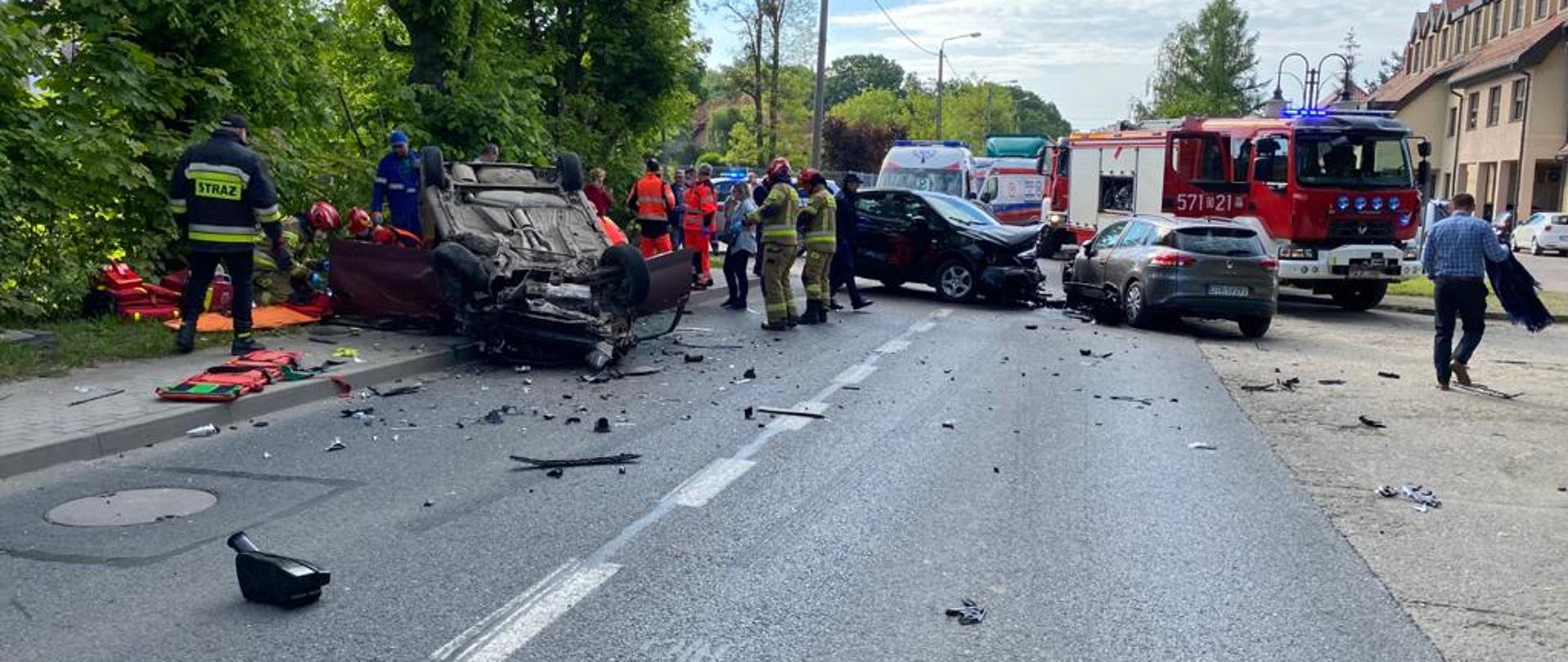 Po lewej stronie samochód na dachu, obok dwa rozbite samochody. Wokół pojazdy służb ratunkowych i ratownicy.