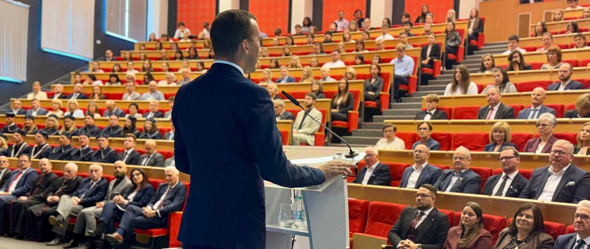 Mężczyzna przemawia do publiczności, stojąc na scenie przy mównicy. 