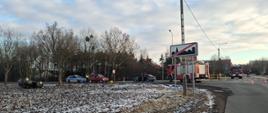 Widok ogólny miejsca kolizji – na poboczu stoi uszkodzony samochód, w tle pojazd straży pożarnej, a w polu obok drogi znajduje się auto przewrócone na dach.