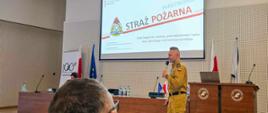 Widok sali konferencyjnej podczas wystąpienia zastępcy lubuskiego komendanta wojewódzkiego PSP. Na ekranie w tle widoczna prezentacja z napisem „Państwowa Straż Pożarna” oraz informacja o Stałym Zespole ds. ochrony przeciwpożarowej lasów województw lubuskiego i zachodniopomorskiego.