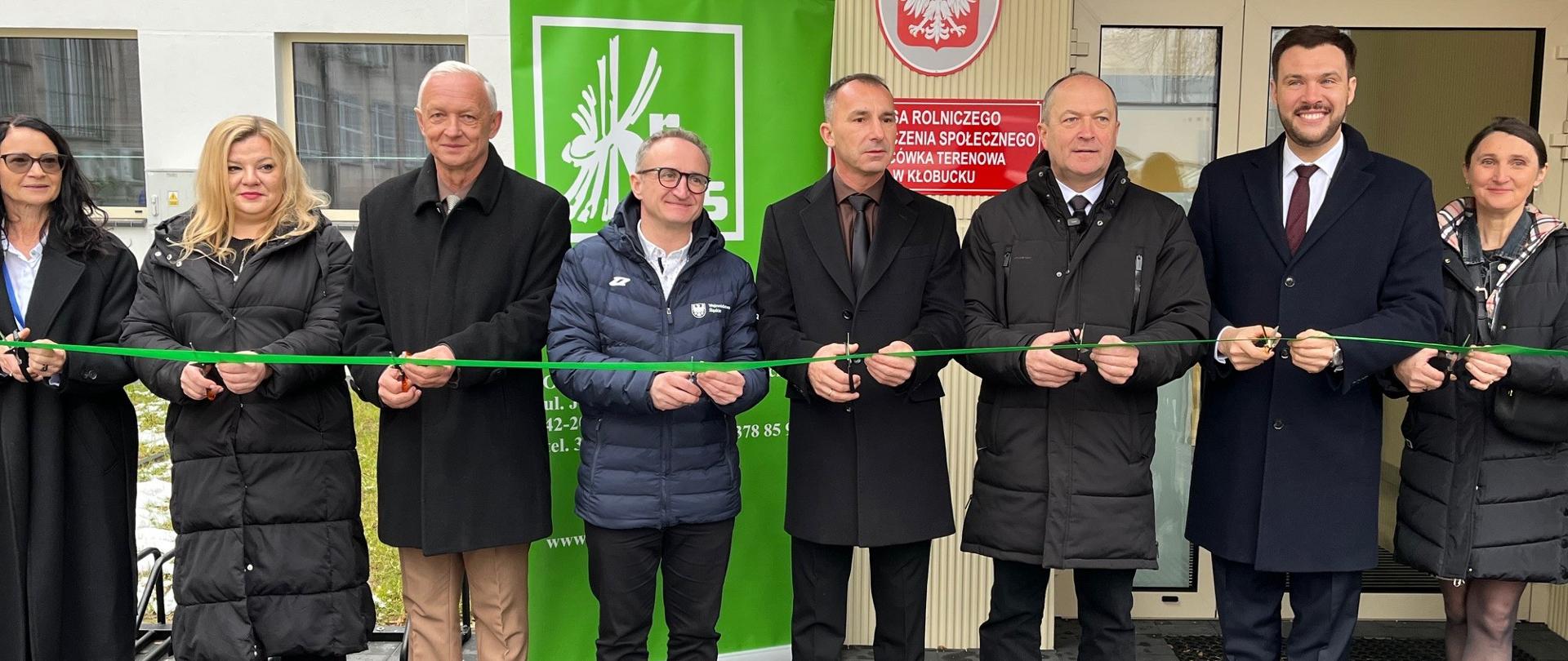 Trzy kobiety i pięciu mężczyzn stoją przed budynkiem i trzymają w rękach zieloną wstęgę i nożyczki. Wszyscy uśmiechają się do zdjęcia.