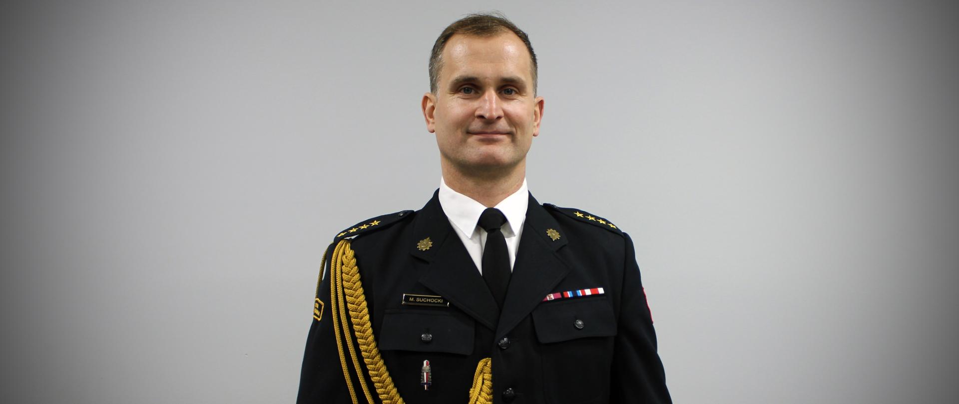 Na zdjęciu widać mężczyznę w eleganckim, galowym mundurze strażackim. Stoi na wprost obiektywu, w wyprostowanej, oficjalnej pozie. Mundur jest ciemny, z pagonami na ramionach, ozdobnym złotym sznurem na prawej stronie oraz kilkoma odznaczeniami i naszywkami. Na piersi znajduje się plakietka z nazwiskiem oraz niewielkie przypinki i barety świadczące o ukończonych szkoleniach lub wyróżnieniach. Mężczyzna ma krótkie, schludnie uczesane włosy i delikatny uśmiech. Tło jest jednolite, jasnoszare, dzięki czemu cała uwaga skupia się na postaci w mundurze