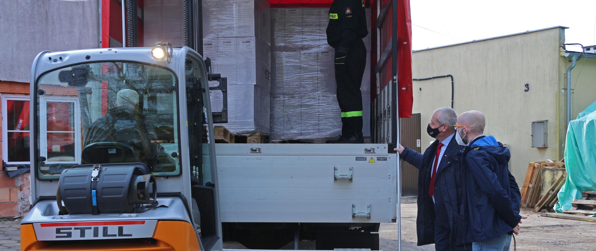 Na zdjęciu na pierwszym planie po lewej stronie widłak, który przewozi paletę z kartonami do stojącego na przeciw widłaka, samochodu ciężarowego. Samochód ma otwartą naczepę. Po prawej stronie, bokiem do obiektywu stoi dwóch mężczyzn. To wojewoda opolski oraz kierownik WCZK. Spoglądają na transport paczek. W tle jasny budynek. 