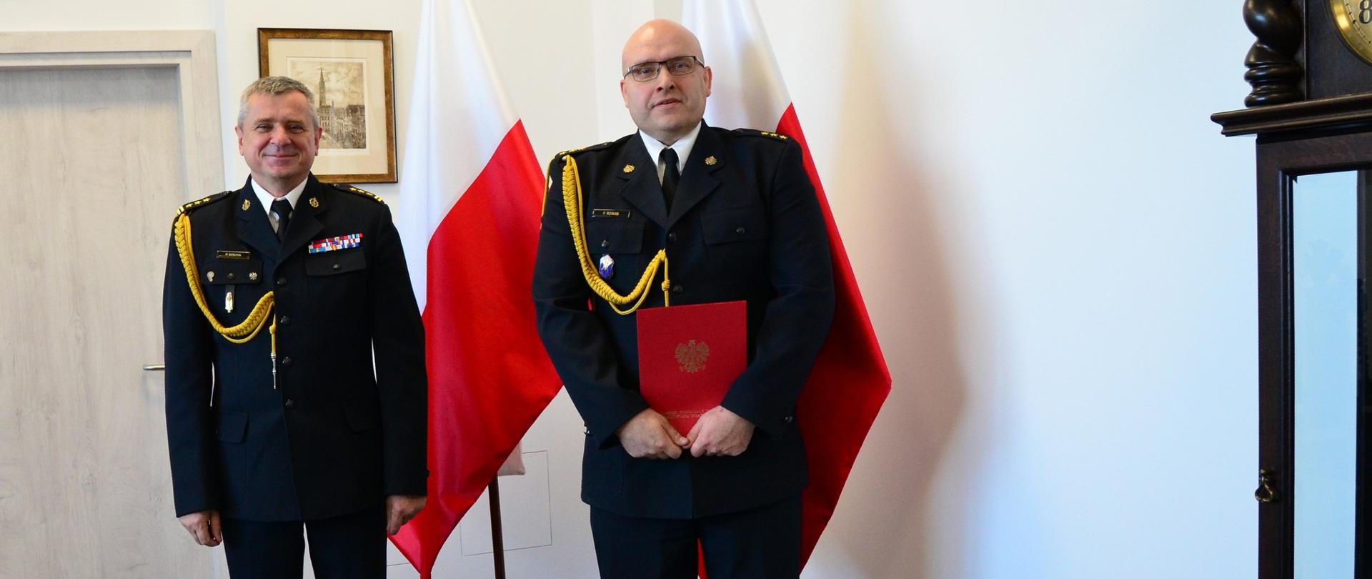 Pomorski komendant wojewódzki Państwowej Straży Pożarnej stoi z funkcjonariuszem trzymającym dokument, strażacy stoją na tle flagi Polski.