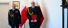 Pomorski komendant wojewódzki Państwowej Straży Pożarnej stoi z funkcjonariuszem trzymającym dokument, strażacy stoją na tle flagi Polski.