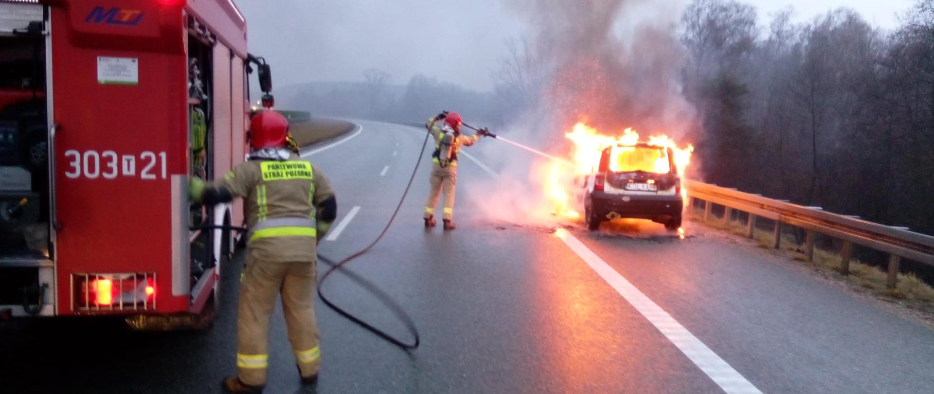 Zdjęcie przedstawia drogę ekspresową. Z lewej strony stoi samochód pożarniczy przy którym strażak nadzoruje pracę autopompy. Drugi strażak podaje wodę na palący się pojazd stojący cały w ogniu po prawej stronie.