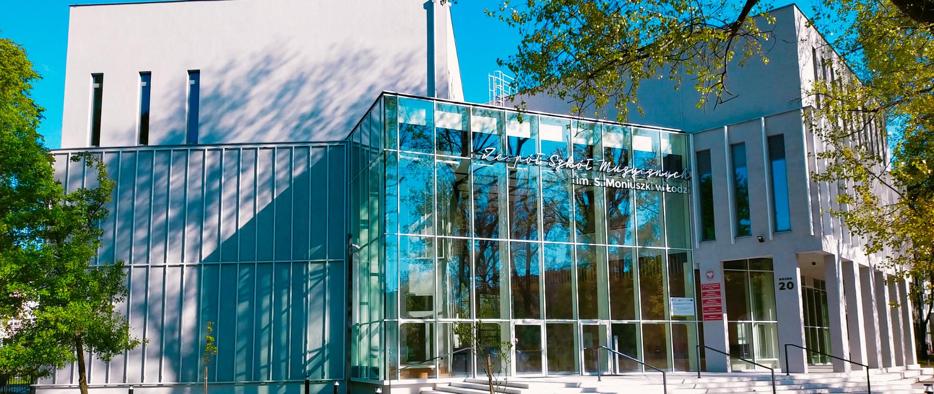 Przeszkolone wejście do nowego budynku szkoły, zaraz przed inauguracją. Na pierwszym planie fragment parku, ławki i kredowe schody. W tle biała elewacja budynku. 