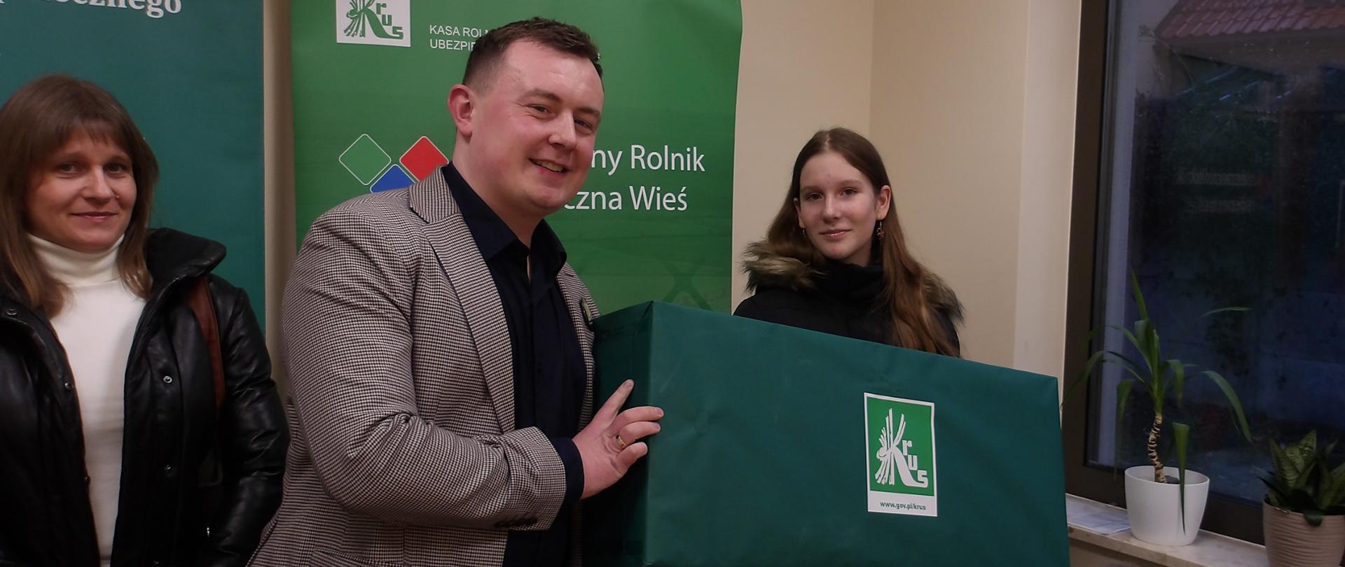 W pomieszczeniu na tle rollupów KRUS mężczyzna wręcza młodej dziewczynie duży zielony karton z logo KRUS, po lewej stronie stoi mama dziewczynki, po prawej mały chłopiec - brat nagrodzonej laureatki.