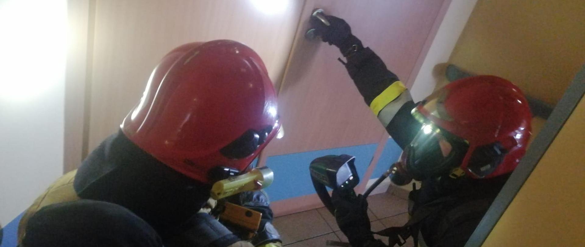 Na zdjęciu widać dwóch ratowników PSP w ubraniach specjalnych, aparatach oddechowych oraz hełmach z kamerą termowizyjną przed wejściem do pomieszczenia w którym jest pożar.