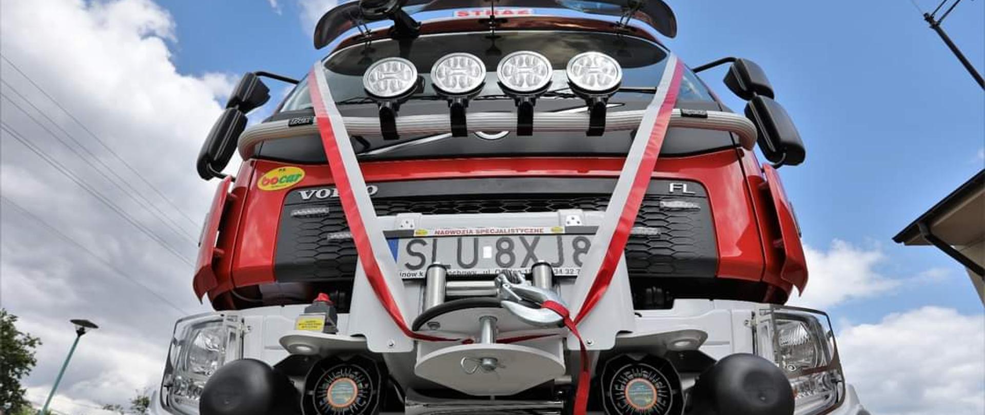 Przepasany biało czerwoną wstęgą przód strażackiego samochodu ratowniczo-gaśniczego.