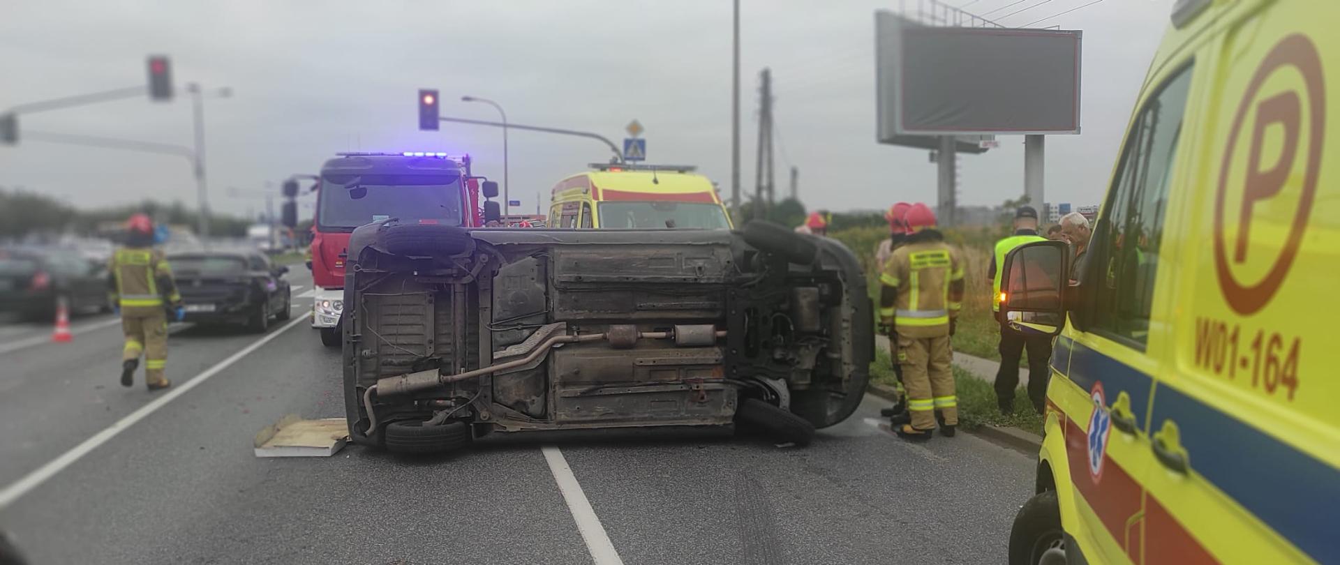 Na zdjęciu widoczny jest uszkodzony samochód leżący na boku, pracujący strażacy. Na zdjęciu widoczny jest samochód straży pożarnej oraz karetki pogotowia