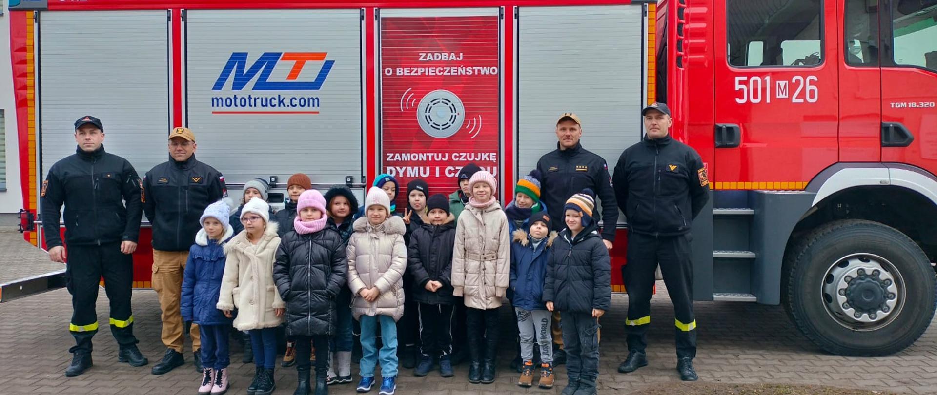 Wspólne zdjęcie dzieci z funkcjonariuszami KP PSP w Makowie Mazowieckim na tle wozu strażackiego. 