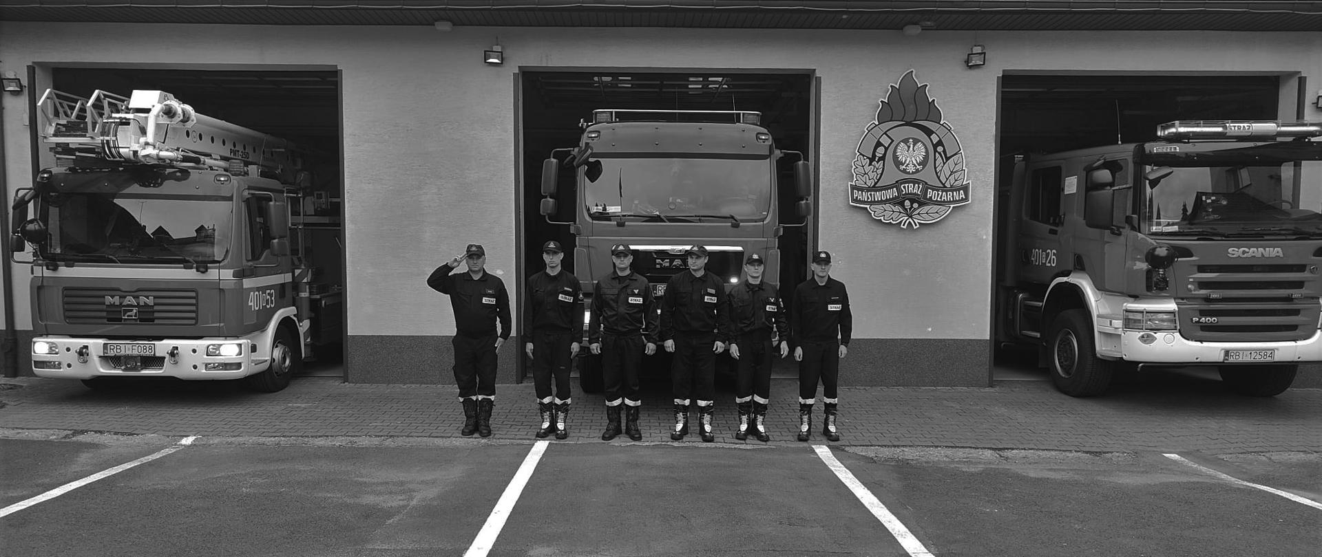 Na zdjęciu widać sześciu strażaków w ubraniach koszarowych na placu zewnętrznym, którzy stoją przed garażami KP PSP w Ustrzykach Dolnych. Dowódca oddaje honor, z tyłu widać pojazdy strażackie które mają włączone sygnały świetlne.