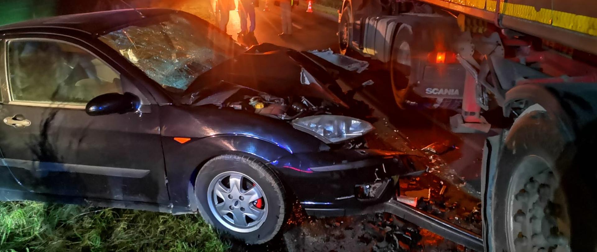 Po lewej stronie widać ciemny samochód osobowy z uszkodzonym przodem, po prawej uszkodzona naczepą samochodu ciężarowego, jest ciemno
