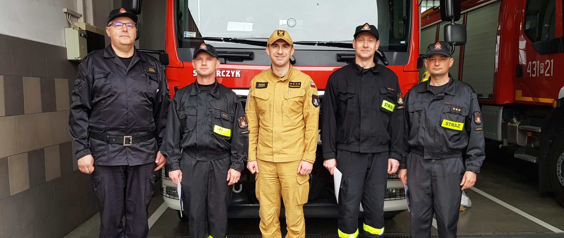 Zdjęcie przedstawia 5 strażaków: komendanta, dowódcę JRG oraz trzech awansowanych strażaków. Stoją oni w szeregu pozując do zdjęcia na tle samochodu ratowniczo-gaśniczego znajdującego się w garażu komendy.