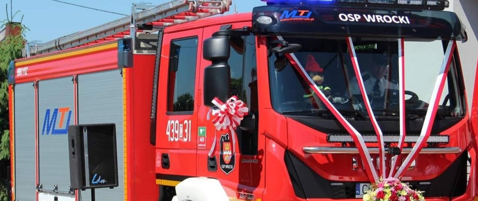 Na zdjęciu widać czerwony wóz ratowniczo-gaśniczy, lusterko oraz przednia szyba są ozdobione biało-czerwonymi wstążkami , przed tablicą rejestracją pojazdu są ułożone kwiaty.