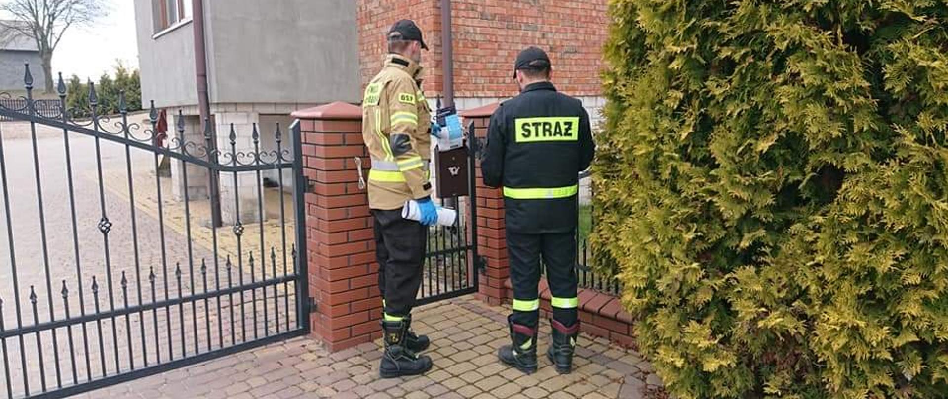 Zdjęcie przedstawia wejście (bramę) do jednego z domostw. Przed brama znajdują się dwaj strażacy w umundurowaniu koszarowym, którzy zostawiają w skrzynce na listy ulotkę informacyjną.