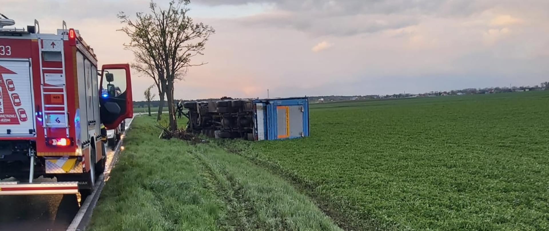 Na zdjęciu samochód ciężarowy poza jezdnia leży na boku oraz zastępy straży pożarnej 