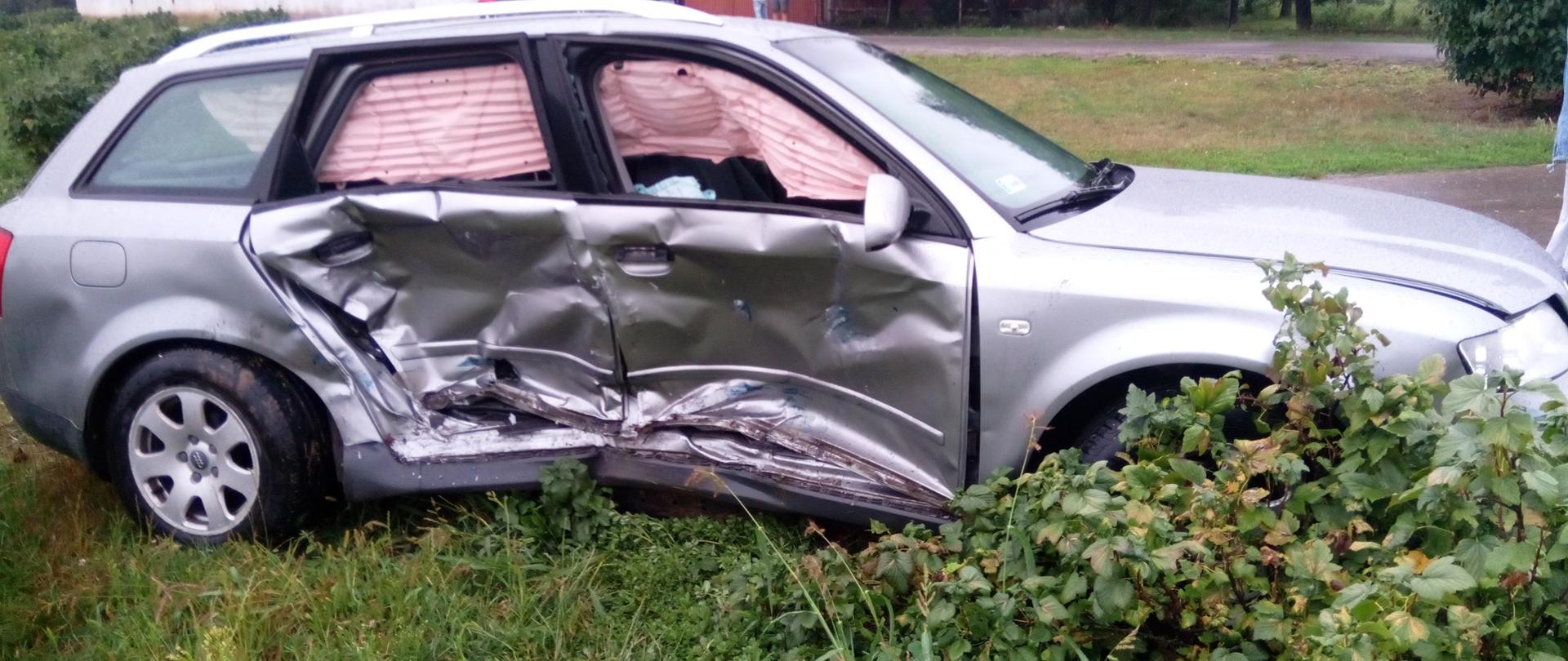 Zdjęcie przedstawia uszkodzony samochód osobowy stojący na poboczu drogi.