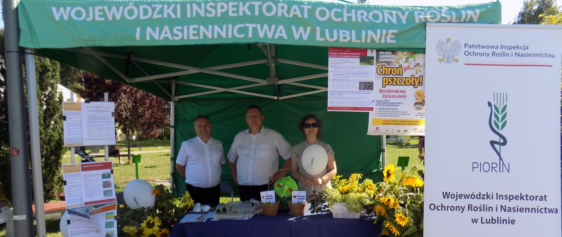 Dwóch mężczyzn w białej koszuli oraz kobieta trzymająca balonik stoją na stoisku pod namiotem. Przed nimi stolik> Na stoliku różne przedmioty i żółte kwiaty. Po bokach stojaki reklamowe.