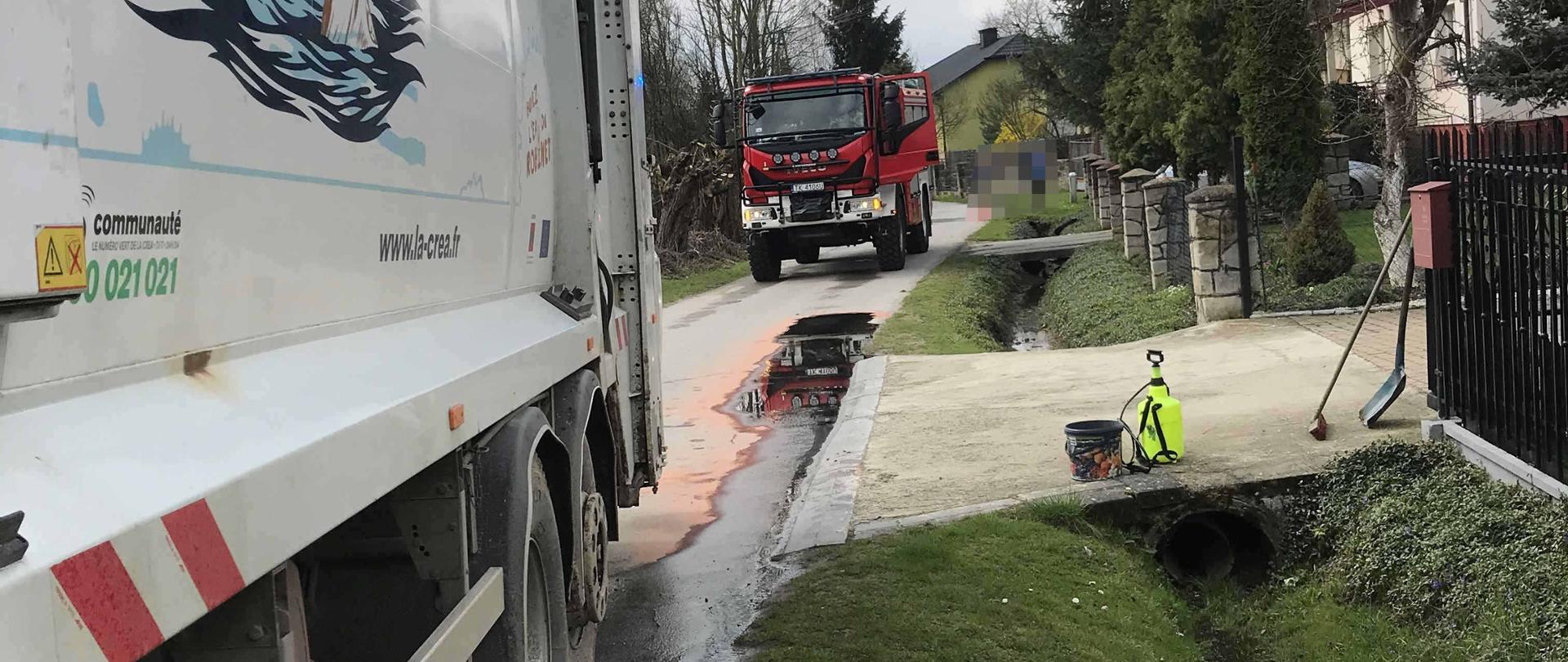 Zdjęcie przedstawia samochód -śmieciarkę oraz rozlany olej hydrauliczny, dalej widać samochód pożarniczy, na podjeździe do posesji widać opryskiwacz ręczny z neutralizatorem , sorbent oraz szuflę i miotłę