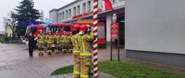Trzech strażaków salutujących i wciągających flagę na maszt. Przed budynkiem Straży Pożarnej stoją samochody oraz inni strażacy.