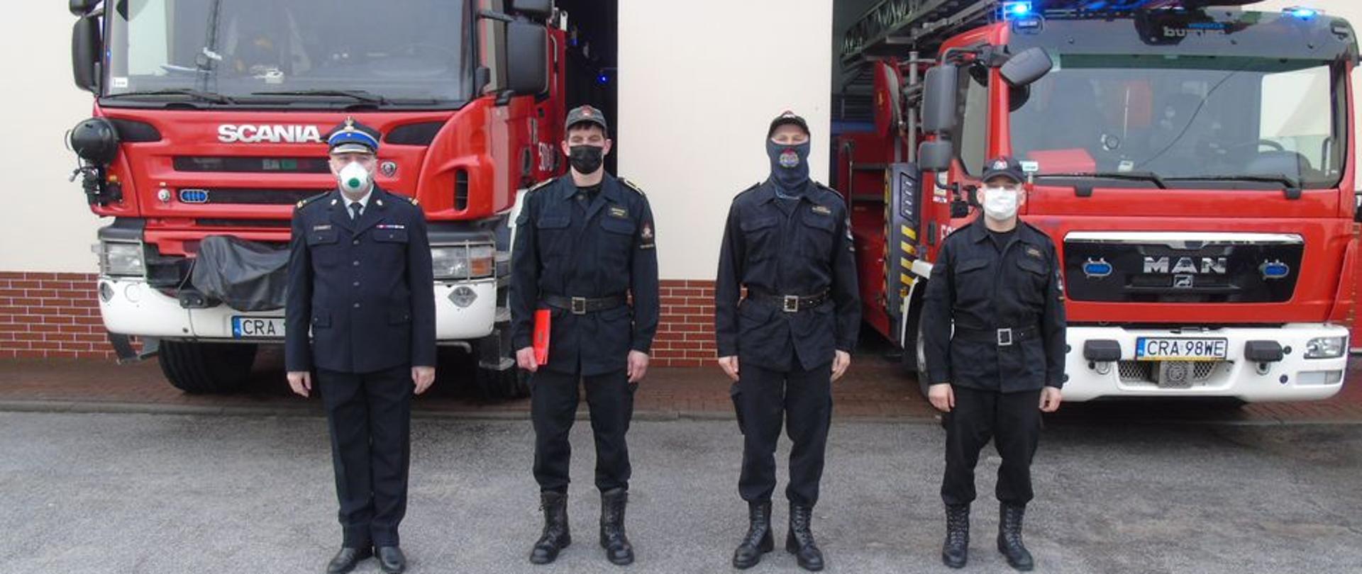 Na pierwszym planie stoi czterech strażaków. Wszyscy w maseczka jeden strażak po lewej stronie ubrany w mundur wyjściowy ( gabardyna), pozostali strażacy ubrani w mundury dowódczo-sztabowe. W tle wydać dwa czerwone pojazdy pożarnicze, ustawione przodem (widoczne kabiny załogi).
