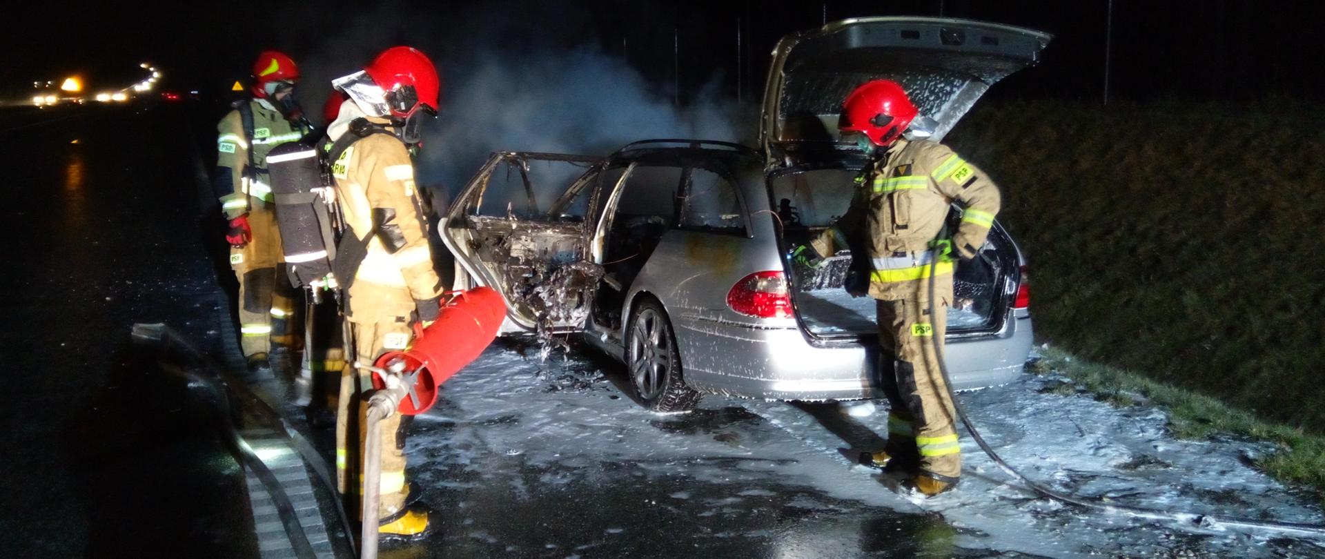 Zdjęcie przedstawia na pierwszym planie strażaków, którzy
zakończyli gaszenie pożaru samochodu. Za nimi znajduje się częściowo
spalony samochód mercedes. Wydobywa się z niego lekki biały dym.
