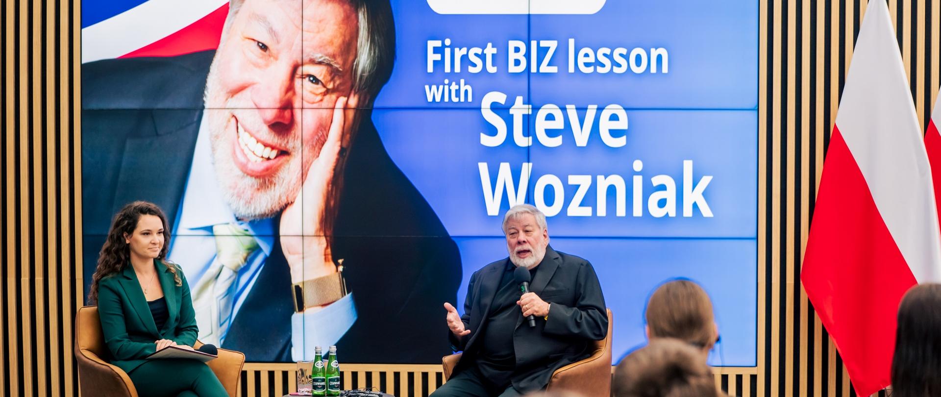Z przodu sali pod wielkim ekranem z napisem First BIZ lesson with Steve Wozniak siedzi Steve Wozniak i mówi do trzymanego w ręku mikrofonu, obok niego kobieta w zielonym garniturze, przed nimi na sali siedzą ludzie.