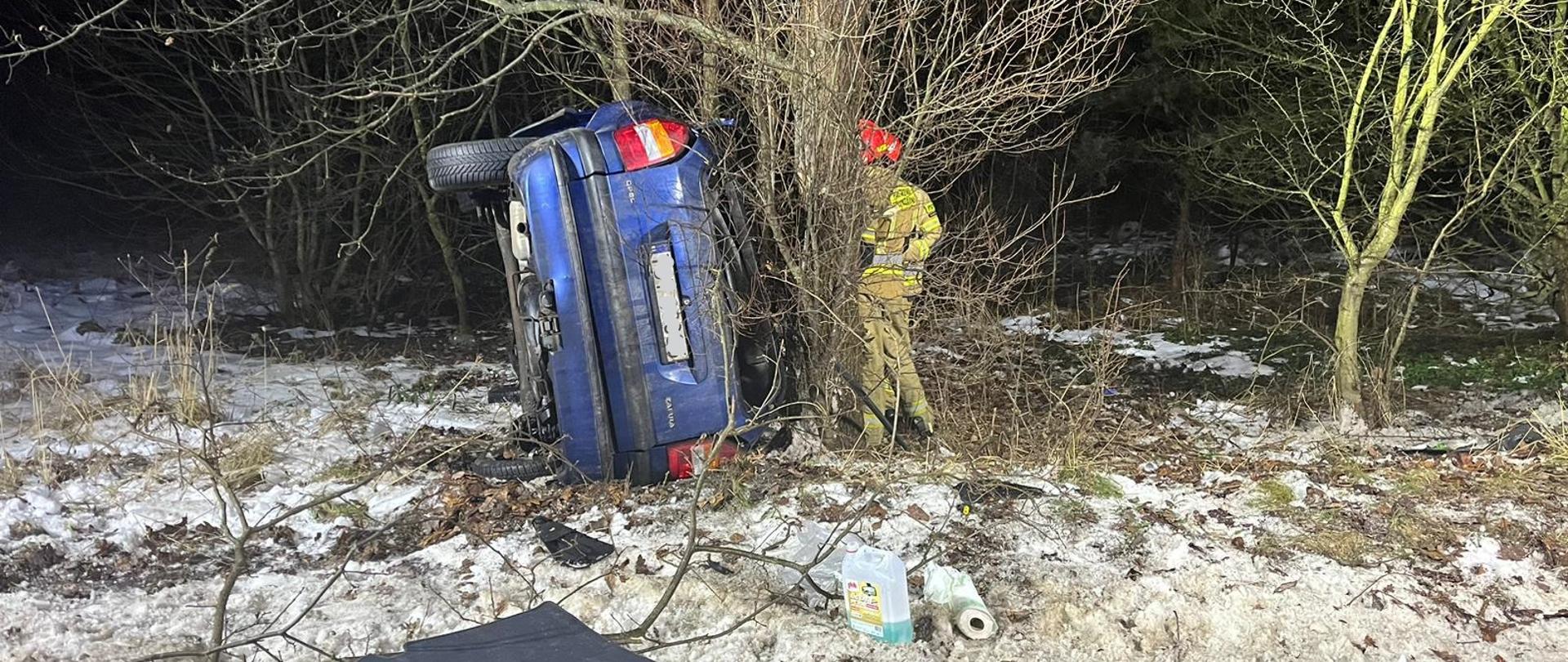 Samochód osobowy leży na prawym boku przy drzewie obok stoi strażak