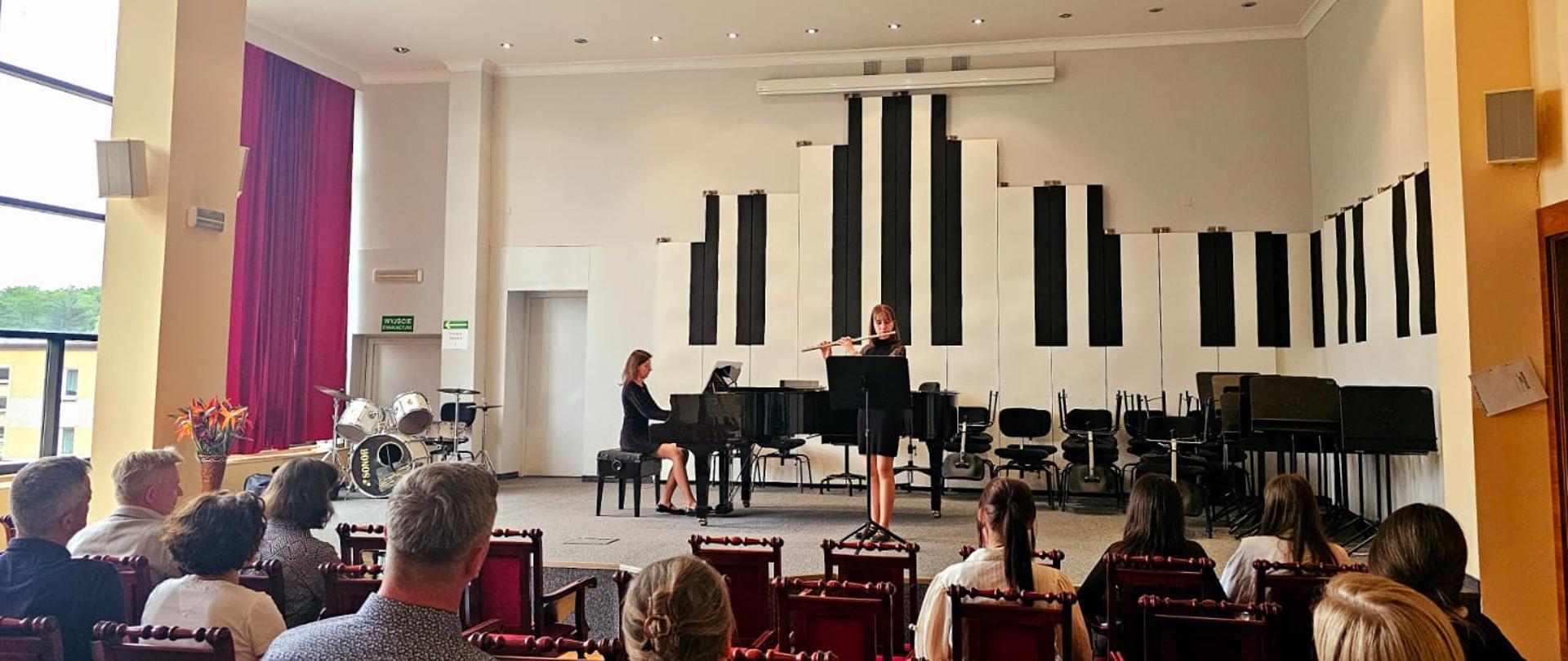 Dziewczyna gra na flecie, kobieta akompaniuje na fortepianie.