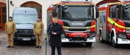 Minister Spraw Wewnętrznych i Administracji przemawiający do zgromadzonych na tle przekazywanych pojazdów pożarniczych. 