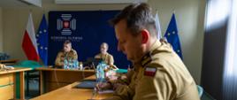 Uczestnicy spotkania w Komendzie Głównej PSP siedzą przy długim stole konferencyjnym. W tle widać flagę Polski, flagę Unii Europejskiej oraz ściankę z logotypem Komendy Głównej PSP. Na stole leżą notatniki, butelki wody i laptopy.