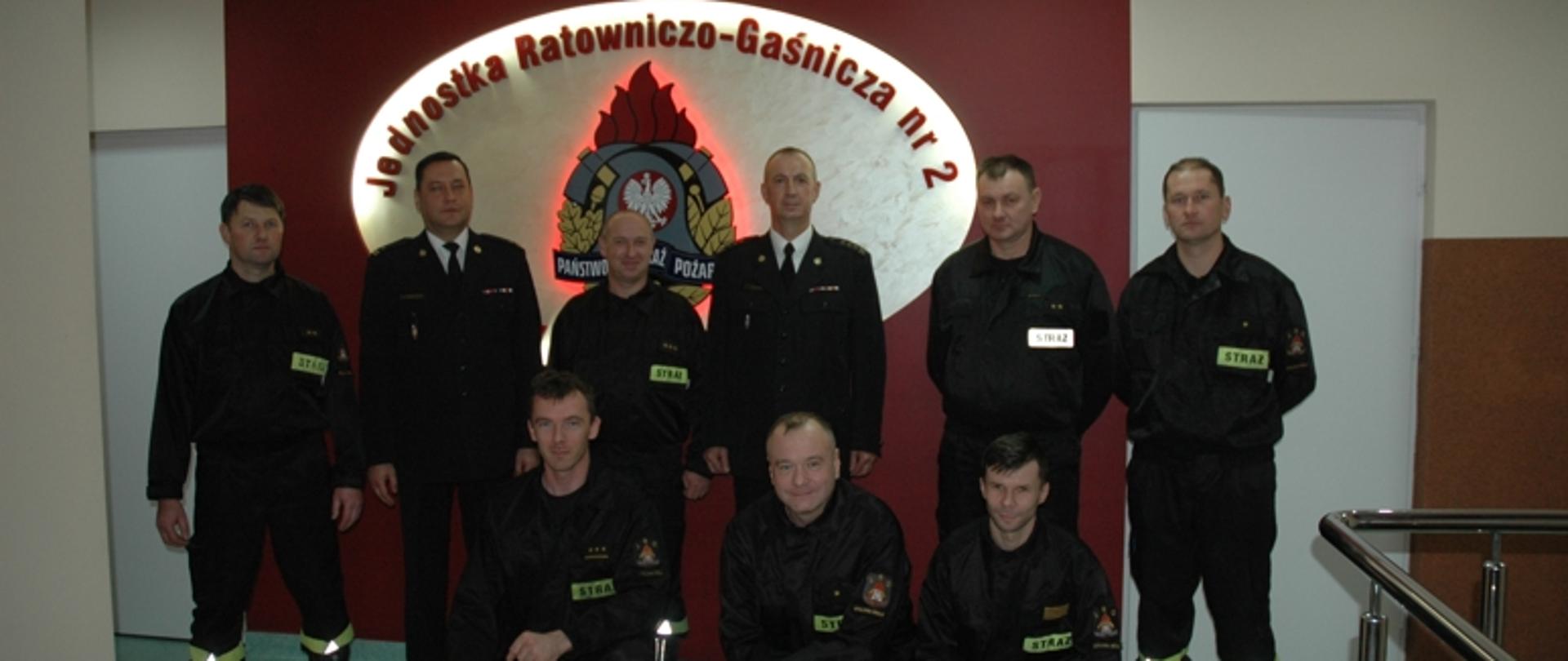 asp. sztab. Mariusz Dziadura z zastępcą komendanta powiatowego PSP w Stalowej Woli st. bryg. Leszkiem Zielińskim, dowódcą Jednostki Ratowniczo-Gaśniczej nr 2 w Stalowej Woli mł. bryg. Marcinem Wróblewskim i obsadą osobową zmiany III z Jednostki Ratowniczo-Gaśniczej nr 2 na tle podświetlanego logo PSP i Jednostki Raotnwiczo-Gaśniczej nr 2 w Stalowej Woli