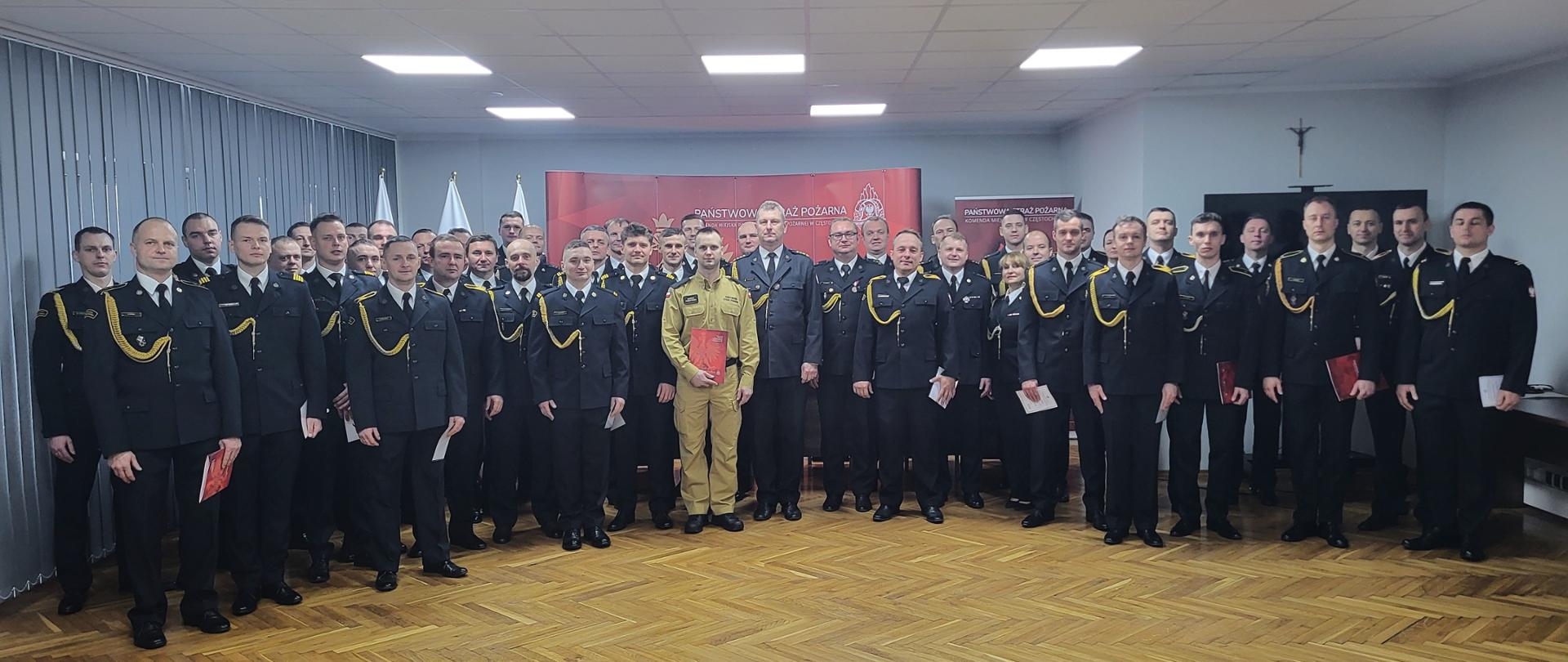 Zdjęcie przedstawia dużą grupę ponad trzydziestu osób, w tym kilka kobiet, stojących razem w pomieszczeniu o drewnianej, parkietowej podłodze. Osoby te są ubrane w galowe mundury Państwowej Straży Pożarnej w kolorze ciemnym. Centralnie z przodu, nieco wysunięty, stoi mężczyzna w jasnym, piaskowym mundurze, trzymający dokumenty w czerwonej oprawie. W tle widoczne są flagi, banery z logo i napisem "PAŃSTWOWA STRAŻ POŻARNA" oraz krzyż na ścianie po prawej stronie.