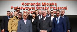  Na zdjęciu grupa kilkunastu osób w większości mężczyzn w garniturach oraz jeden strażak w umundurowaniu – stoi na schodach przed budynkiem z dużym napisem „Komenda Miejska Państwowej Straży Pożarnej”. Pozują do wspólnego, oficjalnego zdjęcia w słoneczny dzień.