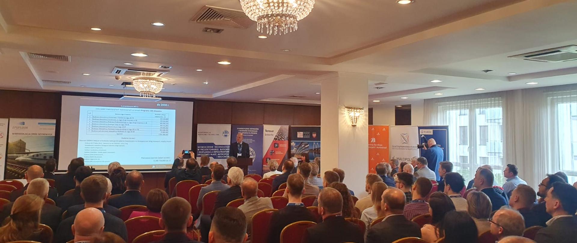 uczestnicy konferencji na pierwszym planie, obserwują wystąpienie, w tle standy, wydarzenie w zamkniętym pomieszczeniu