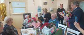 W obecności siedzącej przy stole rodziny, kierownik PT KRUS w Sztumie Elżbieta Akierman wręcza Jubilatce list gratulacyjny obok stoi pracownik placówki z bukietem kwiatów.