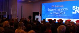 Widok z boku na zaciemnioną wypełnioną siedzącymi ludźmi salę, przed nimi za mównica stoi wiceminister Mrówczyńska i mówi do mikrofonu, za nią na ścianie ekran z napisem Studenci zagraniczni w Polsce 2024.