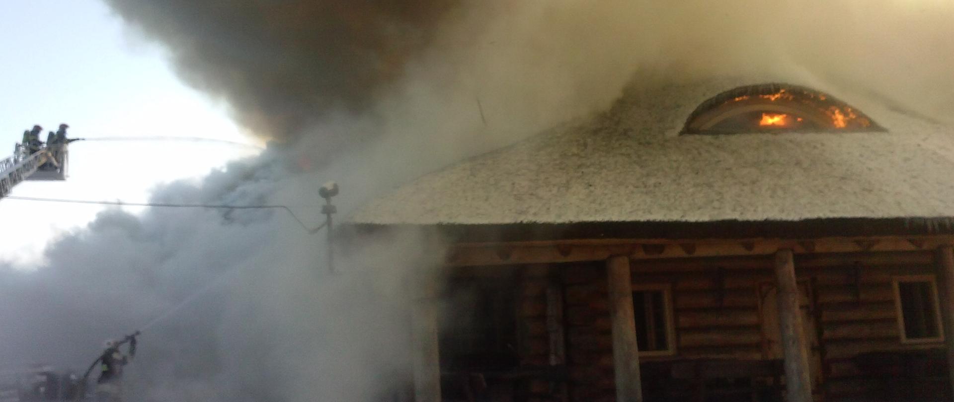 Pożar budynku o konstrukcji drewnianej, dach pokryty strzechą. Nad budynkiem unosi się kolumna szarego dymu. W oknie dachowym widać ogień. Z lewej strony dwóch strażaków, znajdujących się w koszu podnośnika mechanicznego podaje jedne prąd wody na źródło ognia.