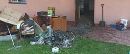 Na zdjęciu widać spalone elementy wyposażenia domu, które są wyniesione na zewnątrz budynku. Obok wejścia do budynku stoi łopata.