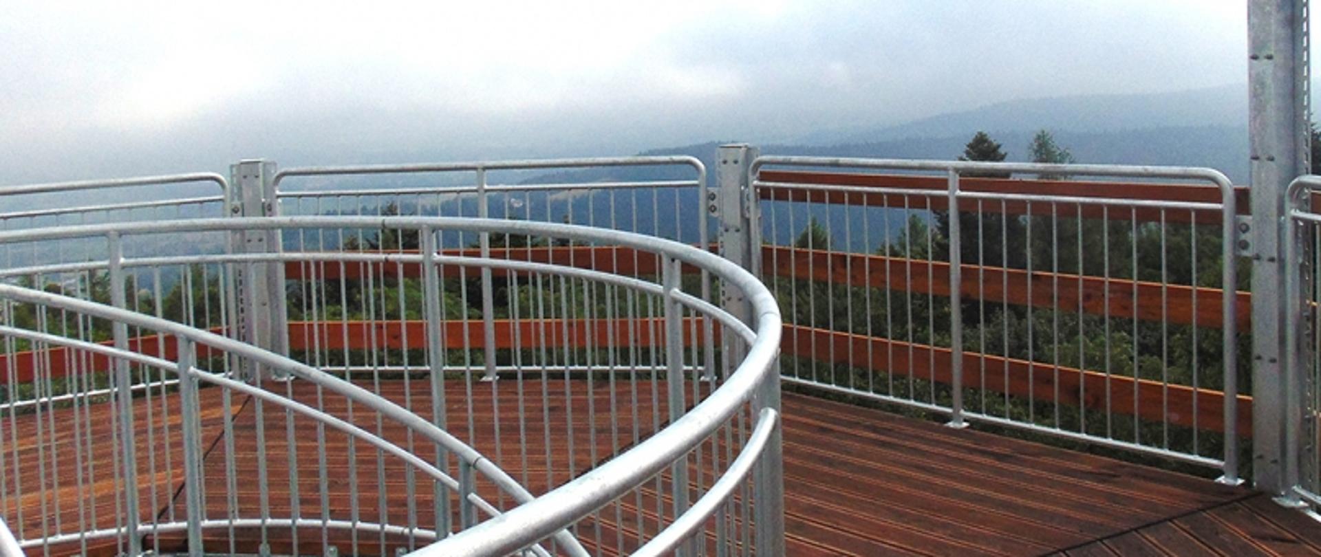 Taras widokowy z widokiem na zalesiony teren i odległe góry pod pochmurnym niebem.