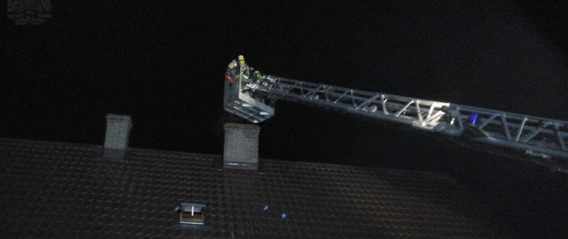 Noc. Strażak w koszu podnośnika wykonuje czynności przy kominie domu jednorodzinnego.