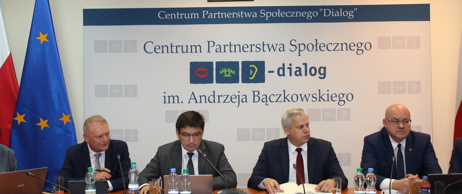Zdjęcie przedstawia stół prezydialny za którym siedzą: Karol Rabenda - Podsekretarz Stanu w Ministerstwie Aktywów Państwowych, Współprzewodniczący Zespołów ze strony rządowej, Pan Krzysztof Kisielewski, Współprzewodniczący Zespołu Trójstronnego ds. Branży Energetycznej ze strony związków zawodowych, Pan Wojciech Ilnicki, Współprzewodniczący Zespołu Trójstronnego ds. Branży Węgla Brunatnego ze strony związków zawodowych oraz Pan Sławomir Wochna, Współprzewodniczący Zespołu Trójstronnego ds. Branży Węgla Brunatnego ze strony pracodawców. W tle siedzących znajduje się baner z napisem Centrum Partnerstwa Społecznego Dialog im. Andrzeja Bączkowskiego. Po lewej stronie zdjęcia stoją dwie flagi: Polska i UE. Nad banerem widnieje Godło Polski.